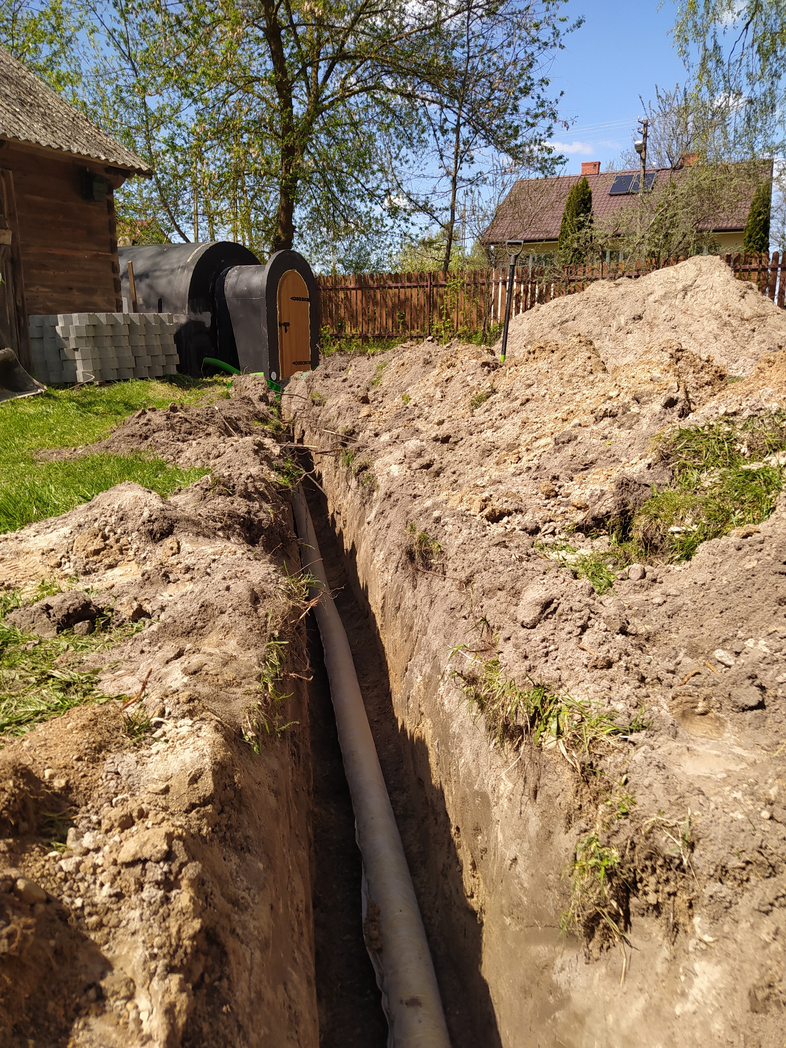 Wąski wykop w ziemi z ułożoną rurą drenażową w folii, pryzma ziemi obok, w tle drewniany budynek gospodarczy i dom z panelami słonecznymi.