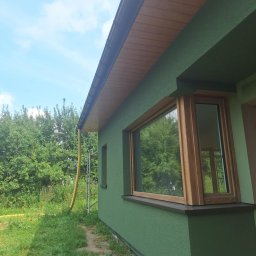 Dawid Drzazga DIDBUD - Ściana budynku z zielonym tynkiem, drewnianą stolarką okienną i rynną, widoczne rusztowanie i trawa
