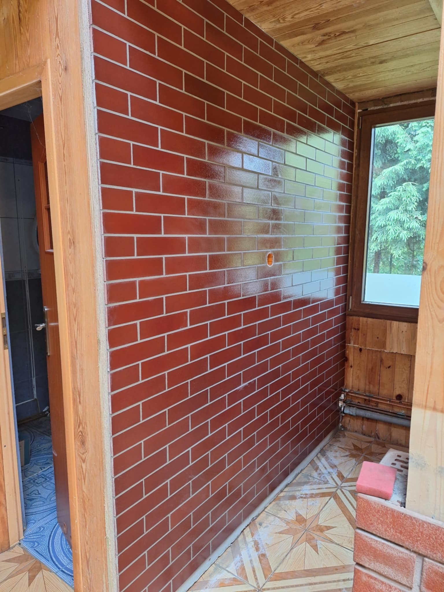 Ściana wyłożona czerwoną cegłą klinkierową z jasną fugą wewnątrz pomieszczenia, widoczne drewniane wykończenia okna i drzwi, oraz fragment podłogi z wzorem gwiazdy.