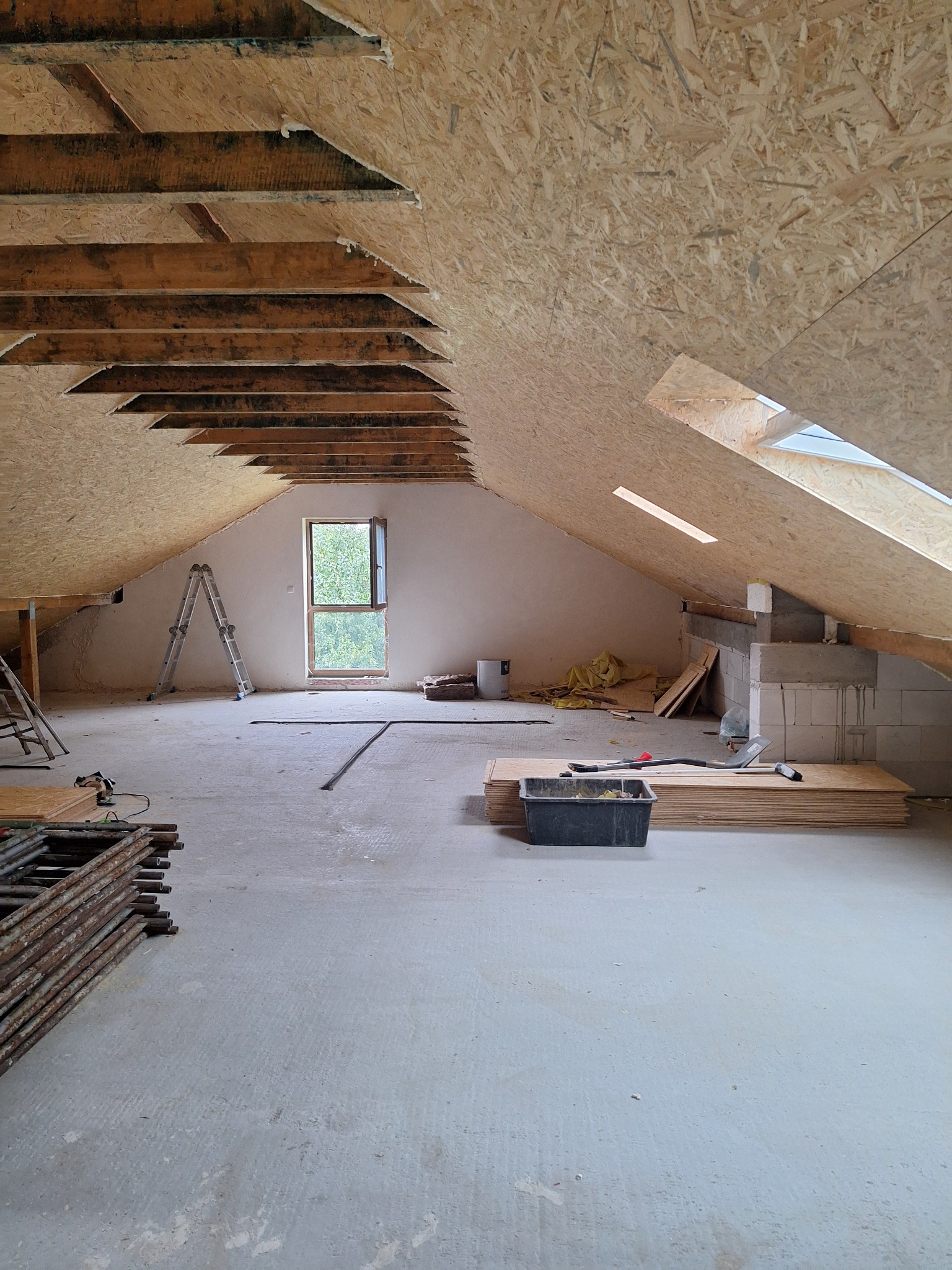 Poddasze w trakcie adaptacji: surowy stan z widoczną konstrukcją dachu, płytami OSB, oknem dachowym oraz rozstawionymi drabinami i narzędziami, przygotowane do dalszych prac.