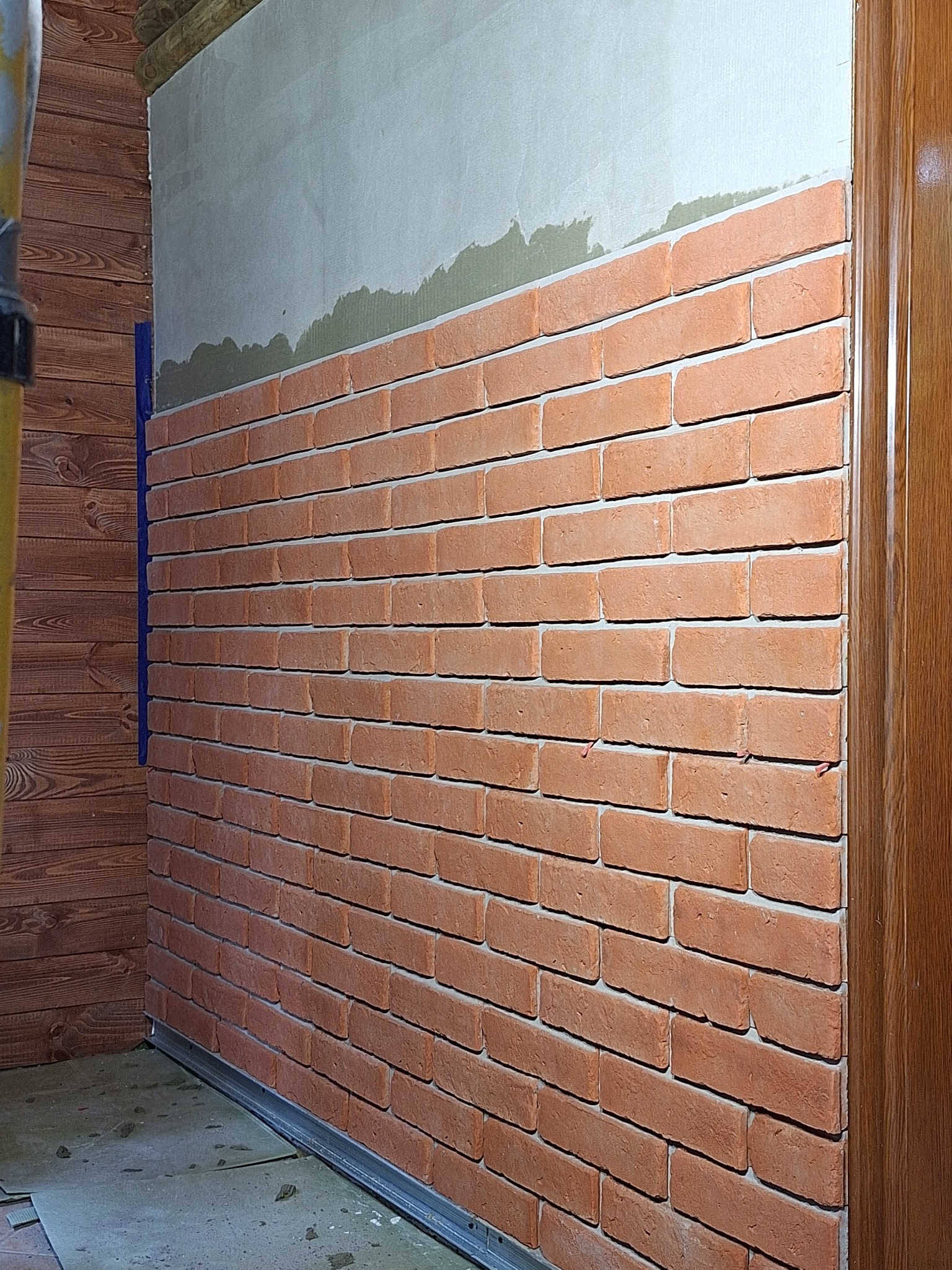 Ściana w trakcie wykańczania z cegieł dekoracyjnych, widoczne niedociągnięcia w górnej części i listwa przypodłogowa, ujęcie z boku.