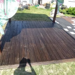 Ciemnobrązowy, drewniany taras z widocznymi słupami konstrukcyjnymi i zasłonami z motywem flamingów, otoczony trawnikiem i elementami ogrodu.