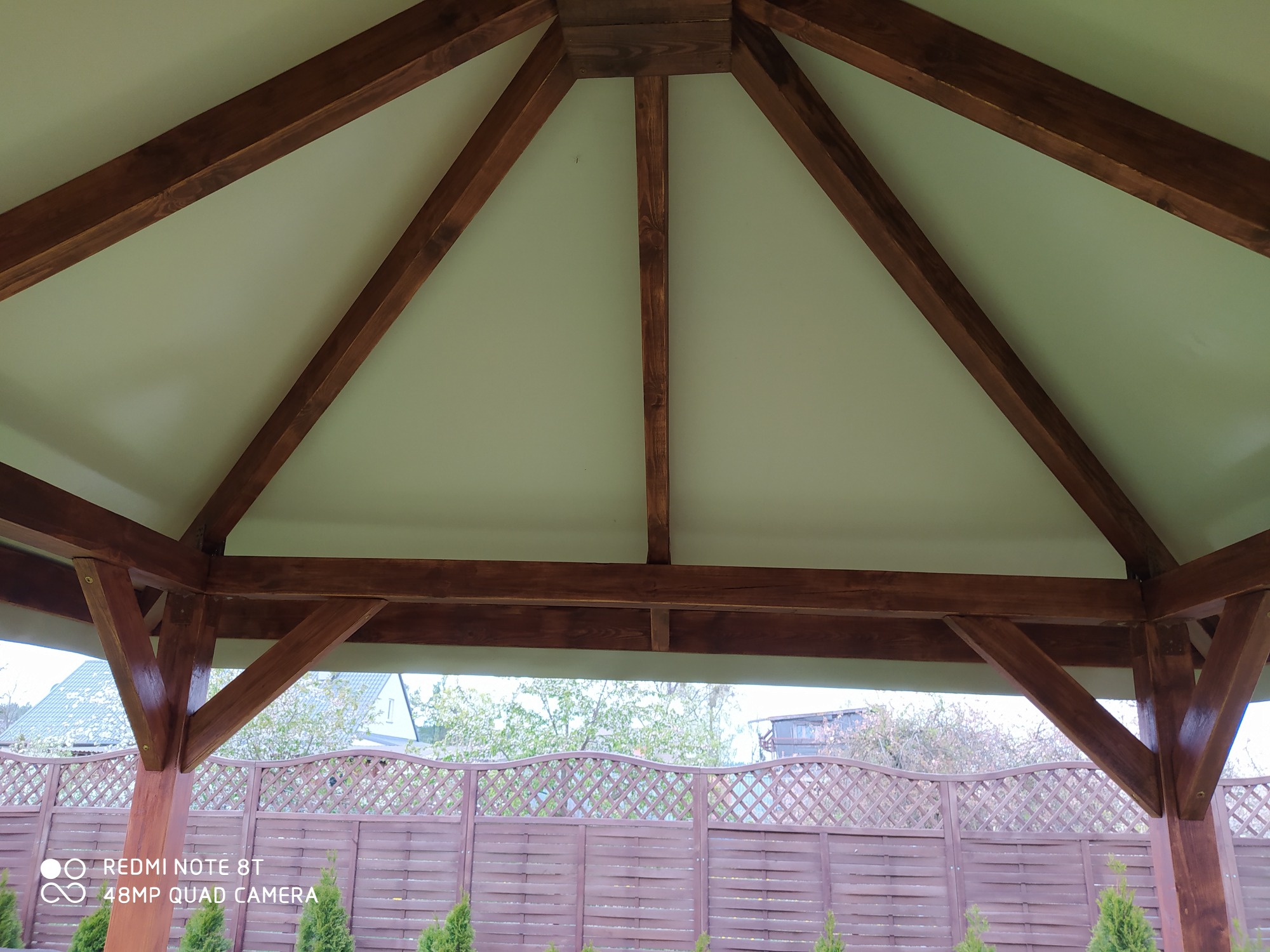 Wnętrze altany z drewnianą konstrukcją dachu, widok z dołu na brązowe belki i jasne poszycie, w tle ogrodzenie i zieleń.