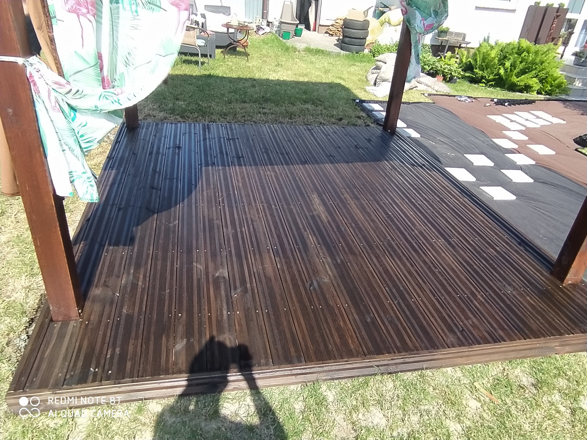 Ciemnobrązowy, drewniany taras z widocznymi słupami konstrukcyjnymi i zasłonami z motywem flamingów, otoczony trawnikiem i elementami ogrodu.