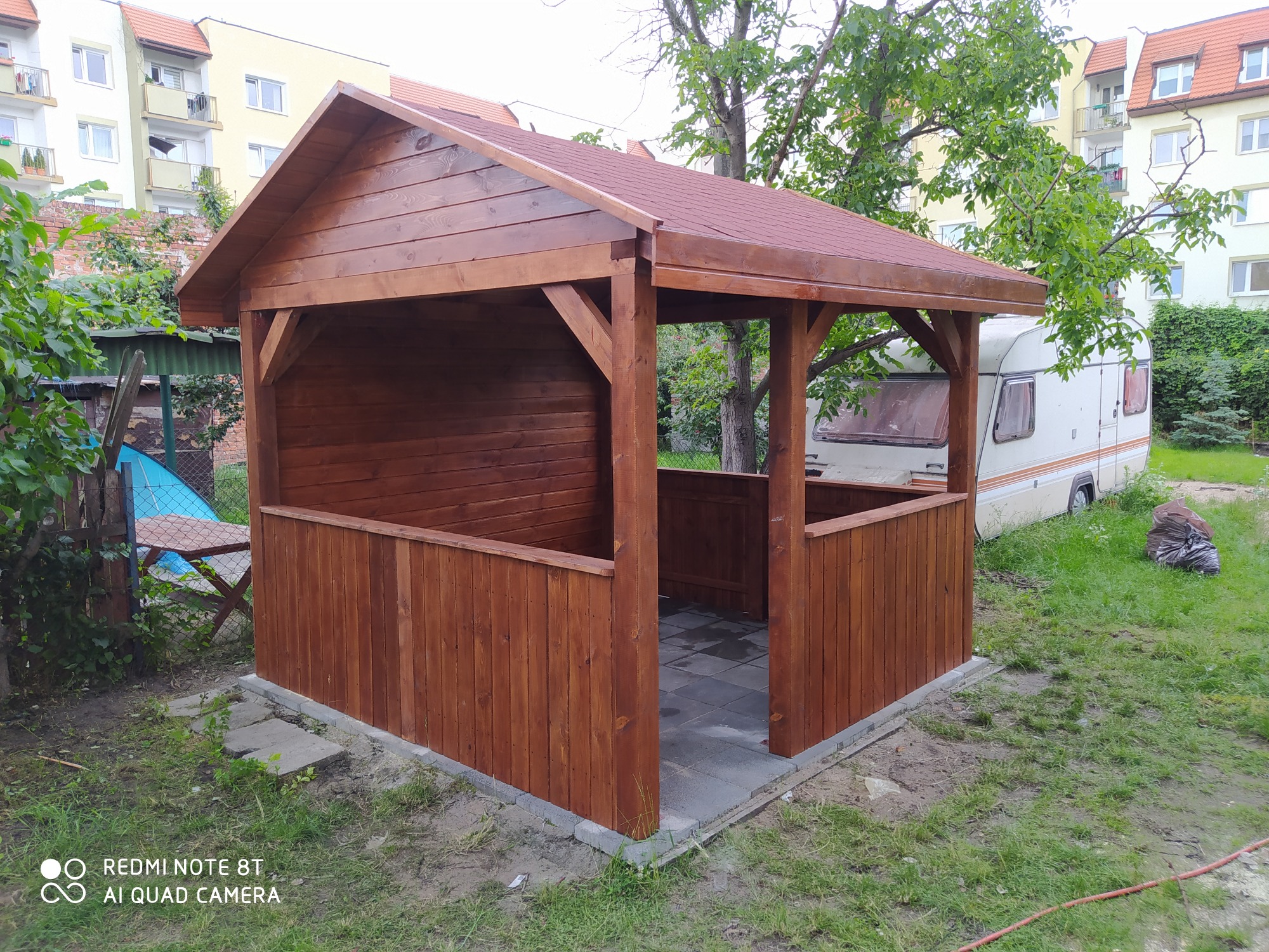 Świeżo postawiona, brązowa konstrukcja z drewna z czerwoną dachówką, otoczona zielenią, z widoczną przyczepą kempingową w tle.