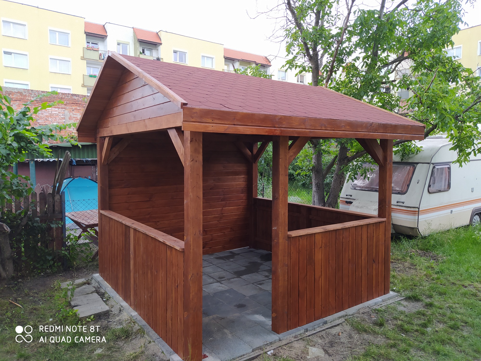 Nowo wybudowana, drewniana altana z czerwoną dachówką na betonowej podłodze, widoczna przyczepa kempingowa w tle.