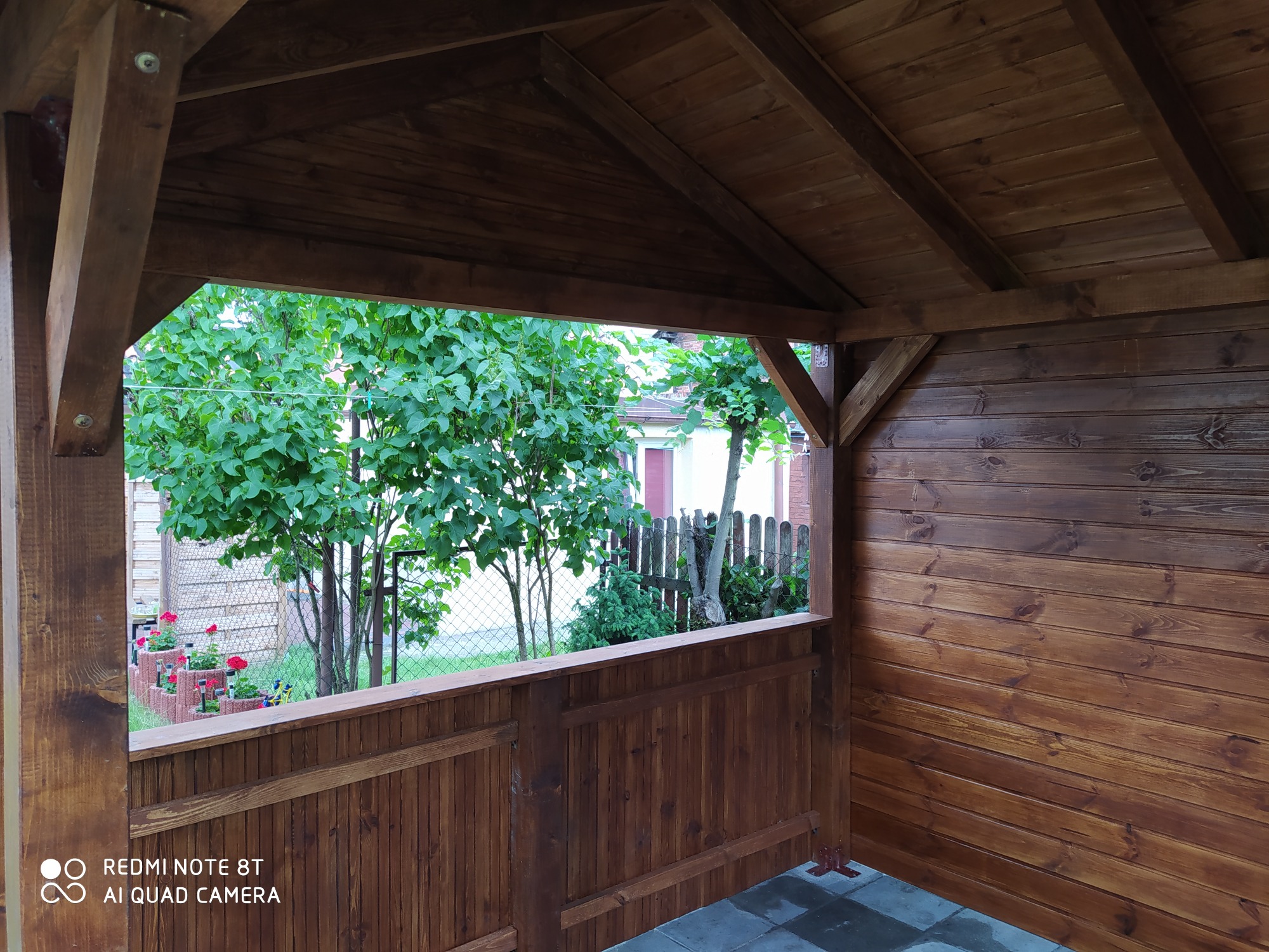 Wnętrze drewnianej altany z widokiem na ogród, widoczne drewniane ściany, podłoga z szarych płyt i roślinność za oknem.