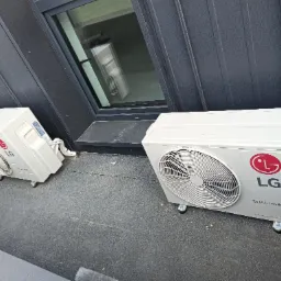 Dwa białe, mobilne klimatyzatory LG Dual Inverter ustawione na szarym, gumowym podłożu przy ciemnej, pionowo żebrowanej ścianie zewnętrznej budynku z oknem.