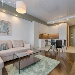 Nowoczesny salon z aneksem kuchennym: beżowa sofa narożna, szklany stolik, dywan w odcieniach zieleni, biała zabudowa kuchenna, drewniana podłoga. Całość w jasnych, stonowanych barwach.