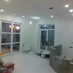 Wnętrze w trakcie prac wykończeniowych: zabezpieczona folią podłoga, przygotowany do obłożenia kominek, białe ściany z widocznymi punktami świetlnymi na suficie i narzędzia w rogu pomieszczenia.