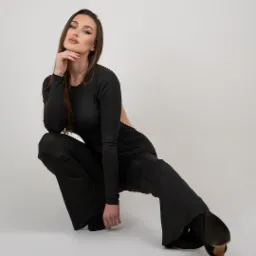 Kobieta w czarnym, dopasowanym stroju pozuje w przysiadzie z ręką pod brodą, eksponując makijaż i buty na obcasie.