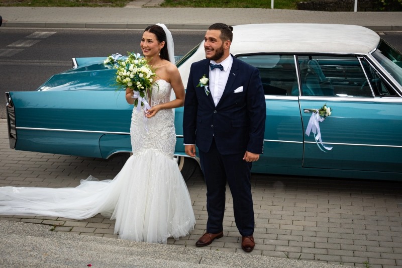 Uśmiechnięta para młoda pozuje przy turkusowym, zabytkowym samochodzie Cadillac z białym dachem, dekorowanym kwiatami i wstążkami.