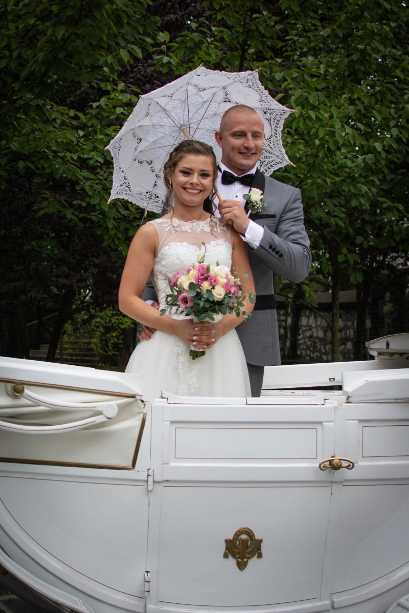 Uśmiechnięta para młoda w białej karocy, panna młoda trzyma bukiet różowych i białych kwiatów, a pan młody trzyma koronkowy parasol nad nimi na tle zieleni.