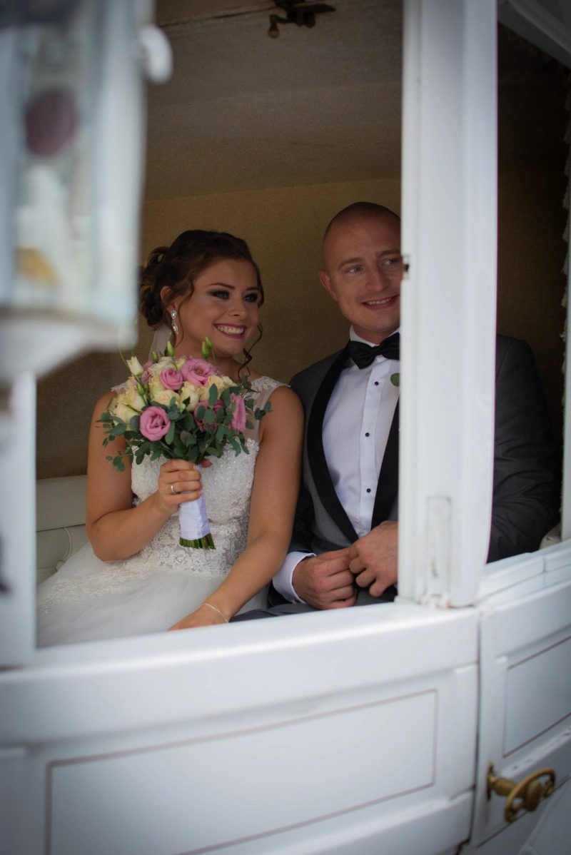 Uśmiechnięta para młoda w karecie, panna młoda trzyma bukiet różowych i białych kwiatów, pan młody w smokingu z muchą, widok przez okno karety.