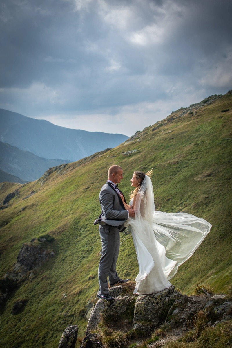Para młoda w strojach ślubnych stoi na skalistym szczycie góry; welon powiewa na wietrze, w tle zielone zbocza i pochmurne niebo.