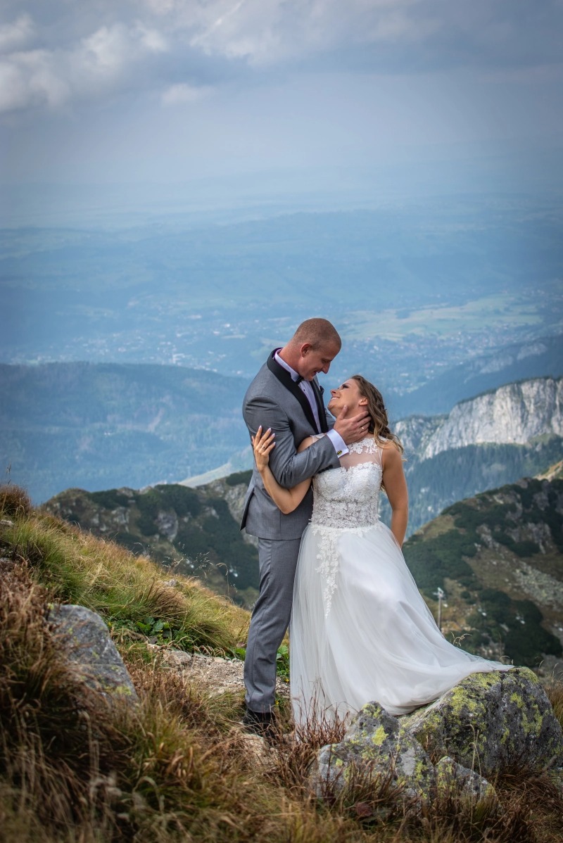 Para młoda, on w szarym garniturze z czarnymi klapami, ona w białej sukni ślubnej z koronkową górą, obejmują się na tle górskiego krajobrazu i pochmurnego nieba.