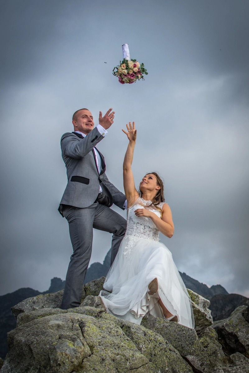 Para młoda w strojach ślubnych na szczycie skalistego wzniesienia rzuca bukiet ślubny w górę, na tle pochmurnego nieba i górskich szczytów.