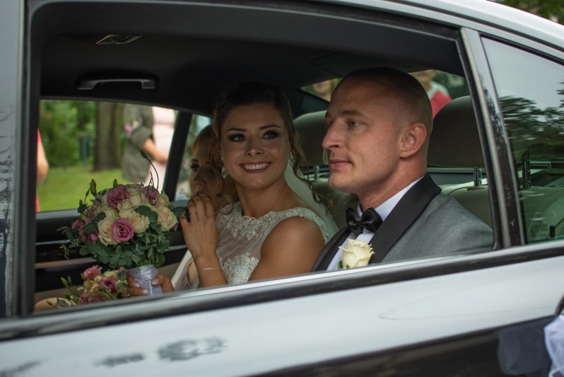 Uśmiechnięta panna młoda w sukni z koronkową górą, siedząca w samochodzie obok pana młodego w szarym smokingu z białą różą w butonierce, trzyma bukiet różowych i białych róż.