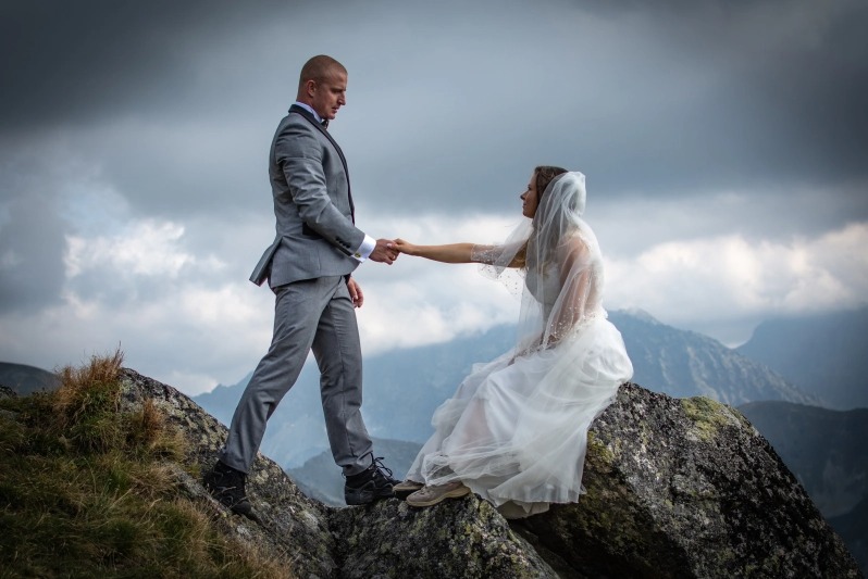 Para młoda w górach: Panna młoda w białej sukni siedzi na skale, a pan młody w szarym garniturze podaje jej rękę na tle pochmurnego nieba i szczytów górskich.