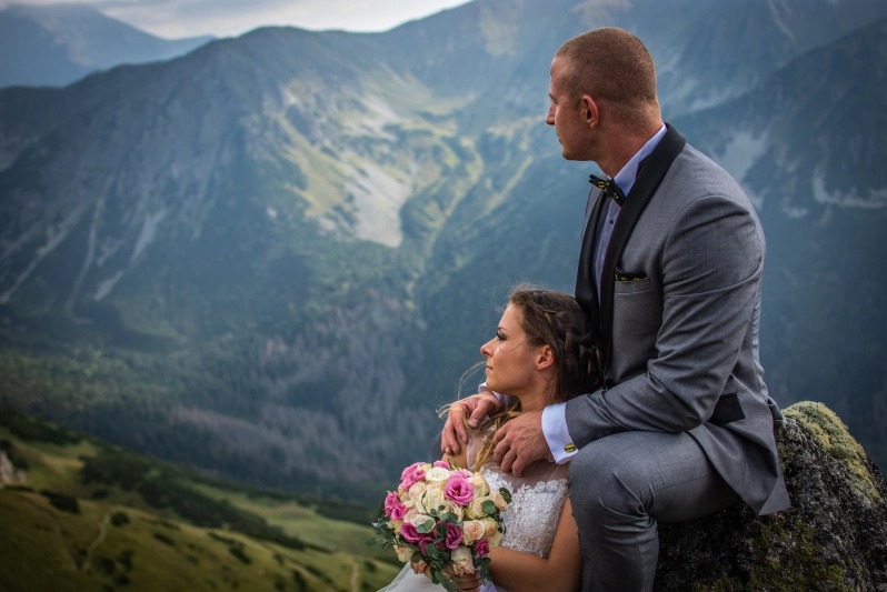 Para młoda w eleganckich strojach siedzi na skale z widokiem na góry; panna młoda trzyma bukiet różowych i białych róż.