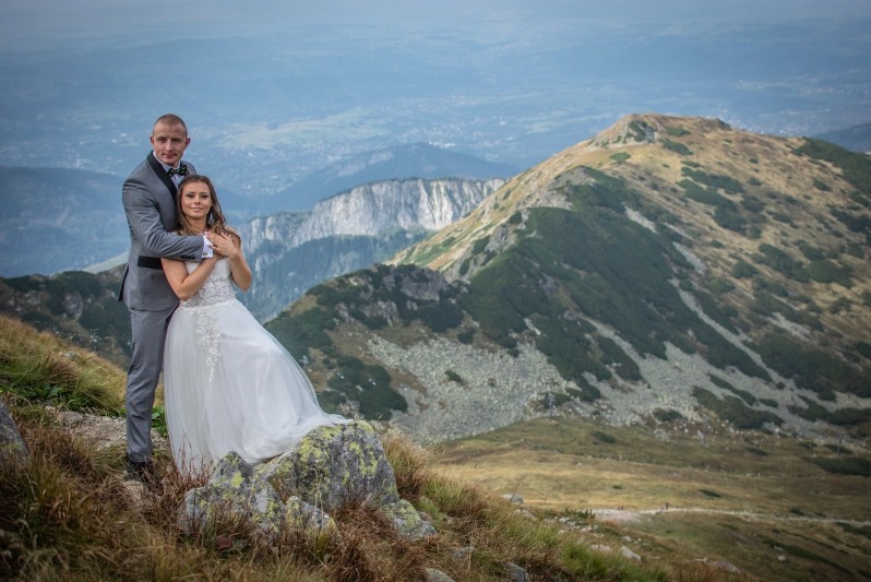 Para młoda w strojach ślubnych pozuje na szczycie góry z rozległym widokiem na dolinę i pasma górskie w tle.