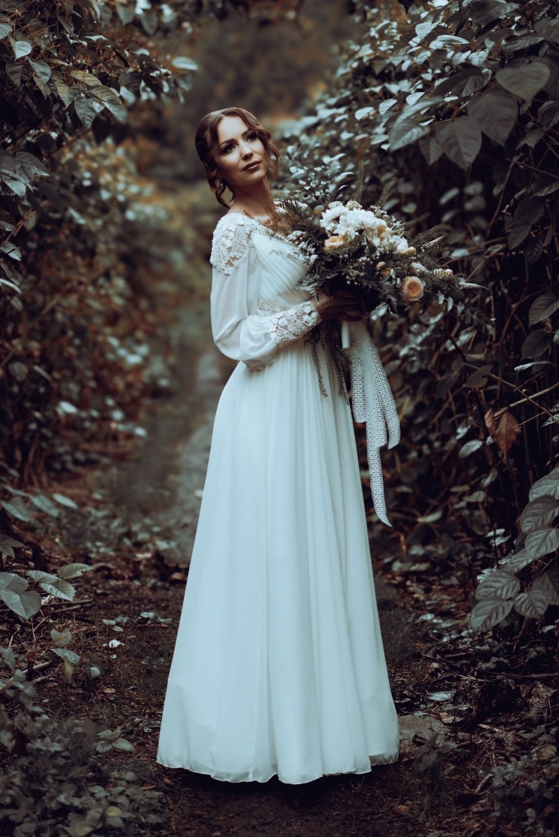 Panna młoda w białej sukni z koronkowymi rękawami, trzymająca bukiet w leśnej scenerii z nisko padającym światłem.
