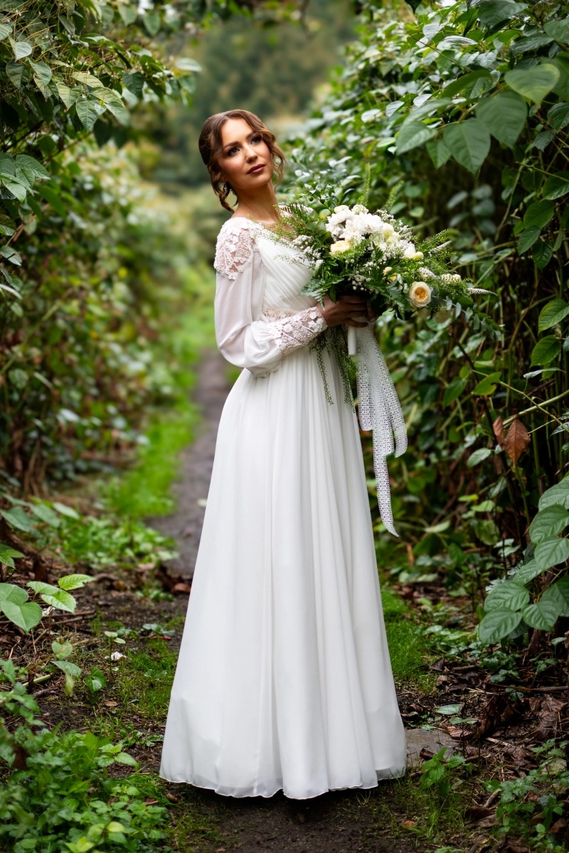 Panna młoda w zwiewnej sukni z koronkowymi rękawami, trzyma bukiet białych kwiatów z zielonymi akcentami, pozuje na leśnej ścieżce otoczonej zielenią.