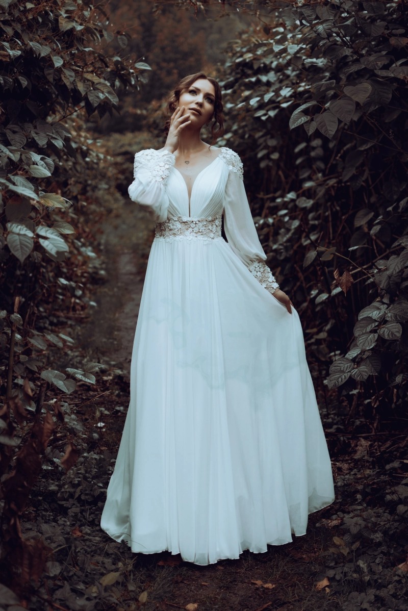 Kobieta w białej, zwiewnej sukni z koronkowymi zdobieniami na rękawach i w pasie, pozująca w zaroślach z dłonią uniesioną do twarzy, delikatny makijaż, romantyczna sesja plenerowa.