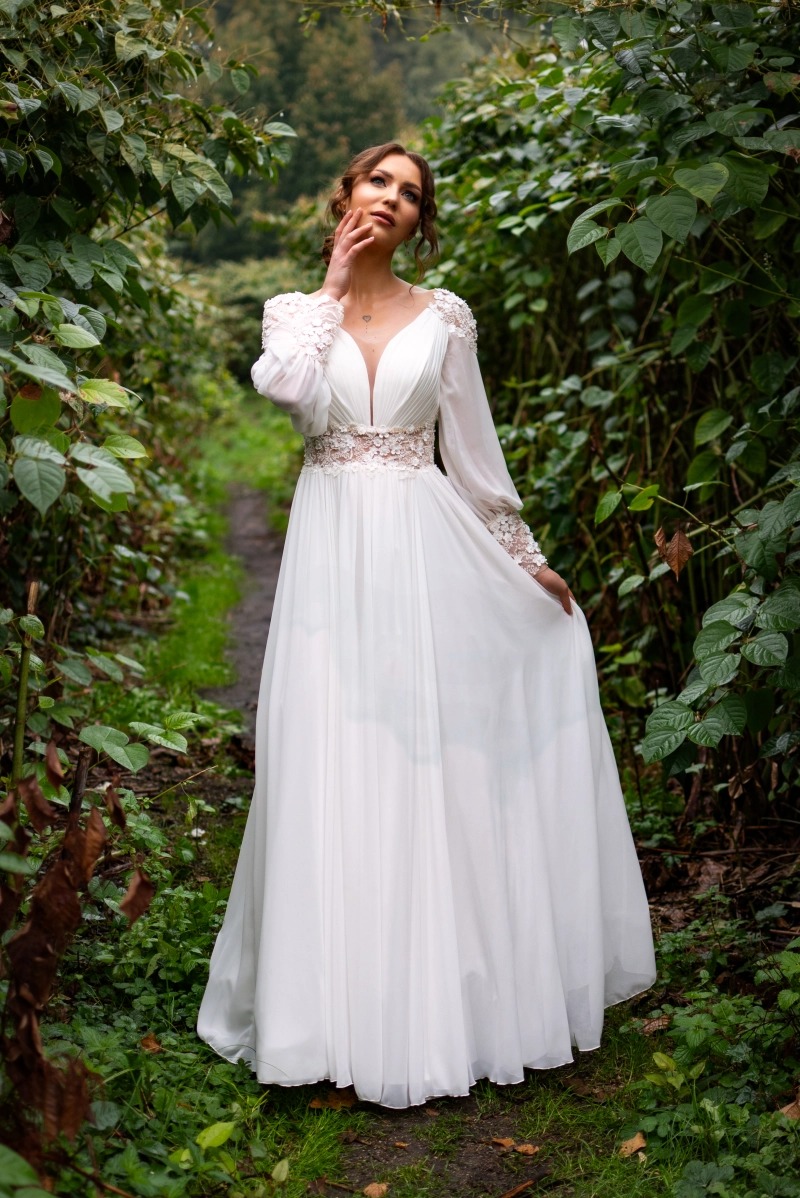 Kobieta w białej, zwiewnej sukni z koronkowymi zdobieniami na rękawach i w pasie, pozuje w zaroślach, trzymając dłoń przy twarzy i spoglądając w górę.