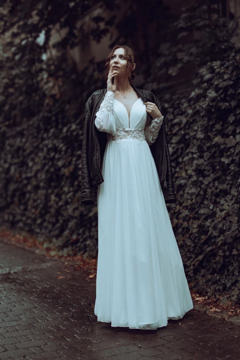 Kobieta w białej sukni z koronkowymi rękawami i skórzanej kurtce na tle ściany z bluszczu, spoglądająca w górę.