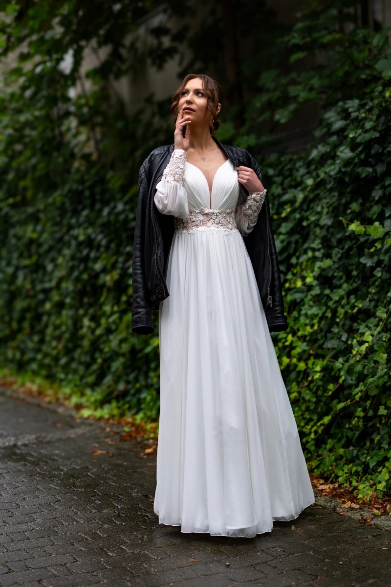 Panna młoda w białej sukni z koronkowymi rękawami i skórzaną kurtką na tle zielonej ściany z bluszczu, zamyślona poza.