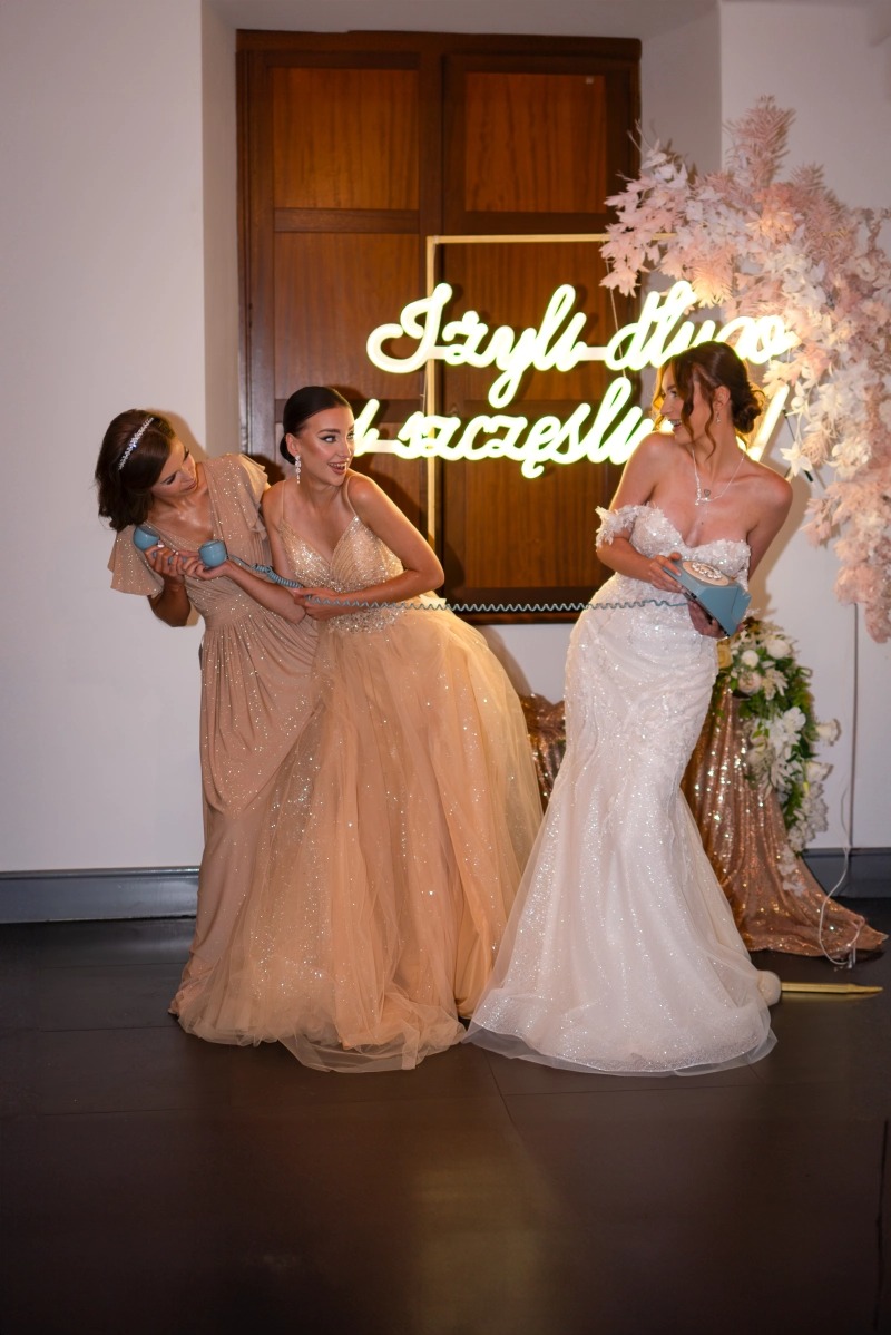Trzy kobiety w eleganckich sukniach pozują z niebieskim, staromodnym telefonem na tle dekoracji ślubnej z neonowym napisem 'I żyli długo i szczęśliwie'.