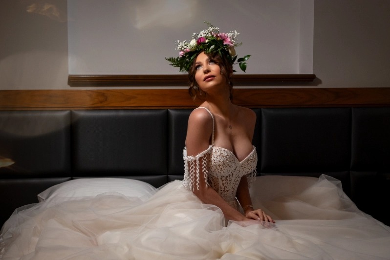 Kobieta w koronkowej sukni z odkrytymi ramionami i wiankiem z kwiatów na głowie, siedząca na łóżku z czarnym skórzanym zagłówkiem, spoglądająca w górę.