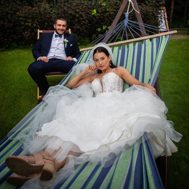 Panna młoda w koronkowej sukni leży w hamaku w ogrodzie, obok siedzi pan młody w granatowym garniturze.