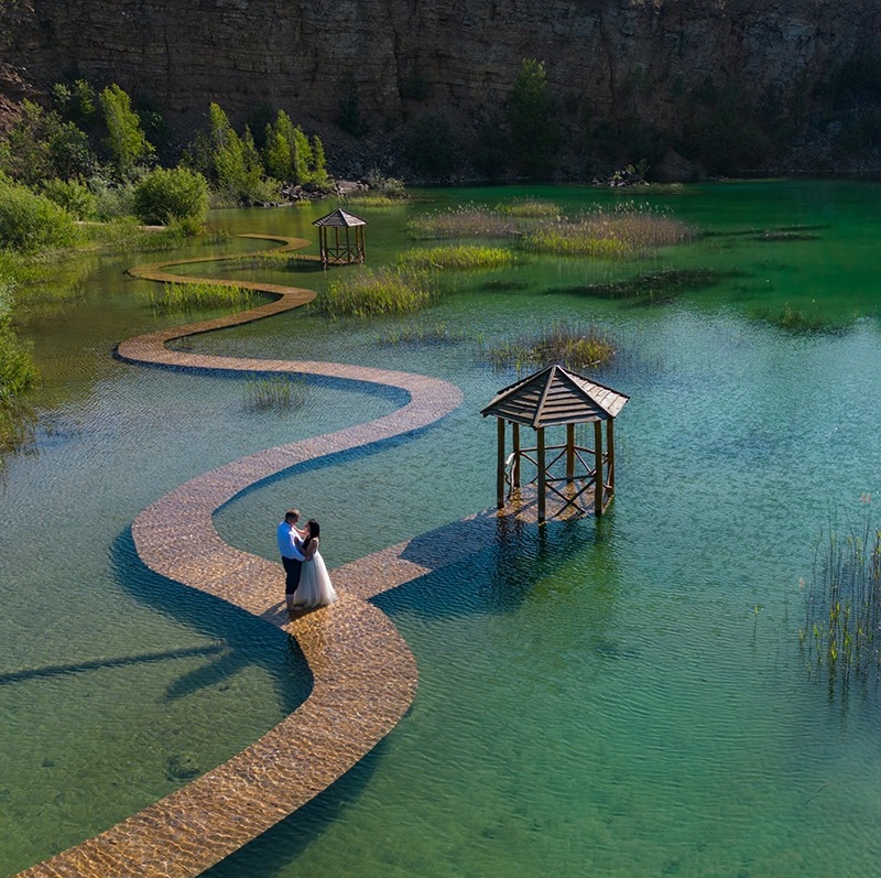 Para młoda obejmuje się na krętej ścieżce wodnej prowadzącej do drewnianej altanki na turkusowym jeziorze, z klifem i drzewami w tle.