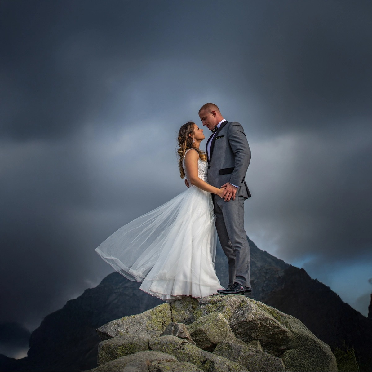 Para młoda, panna młoda w zwiewnej sukni i pan młody w szarym garniturze, stoją na skalistym szczycie góry na tle burzowego nieba, trzymając się za ręce i patrząc sobie w oczy.