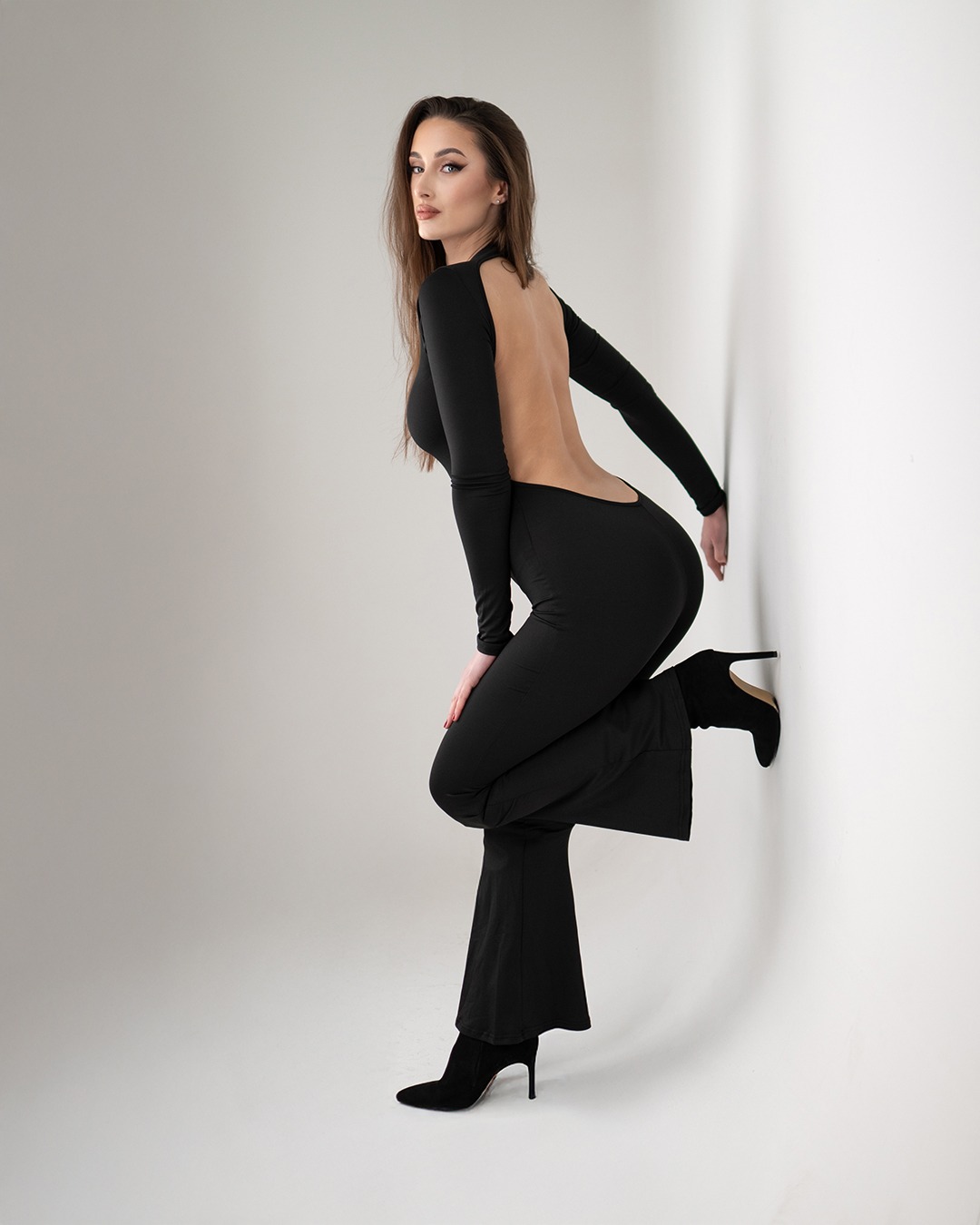 Kobieta w czarnym kombinezonie z odkrytymi plecami, oparta o białą ścianę, pozuje w czarnych butach na obcasie.