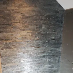 Ściana wykończona dekoracyjną okładziną z wąskich, szarych płytek kamiennych, ułożonych horyzontalnie, z widocznym fragmentem narożnika i sufitu.