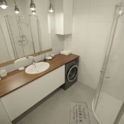 Nowoczesna łazienka z prysznicem typu walk-in, umywalką nablatową na drewnianym blacie, pralką i dywanikiem z napisem 'SHOWER'.
