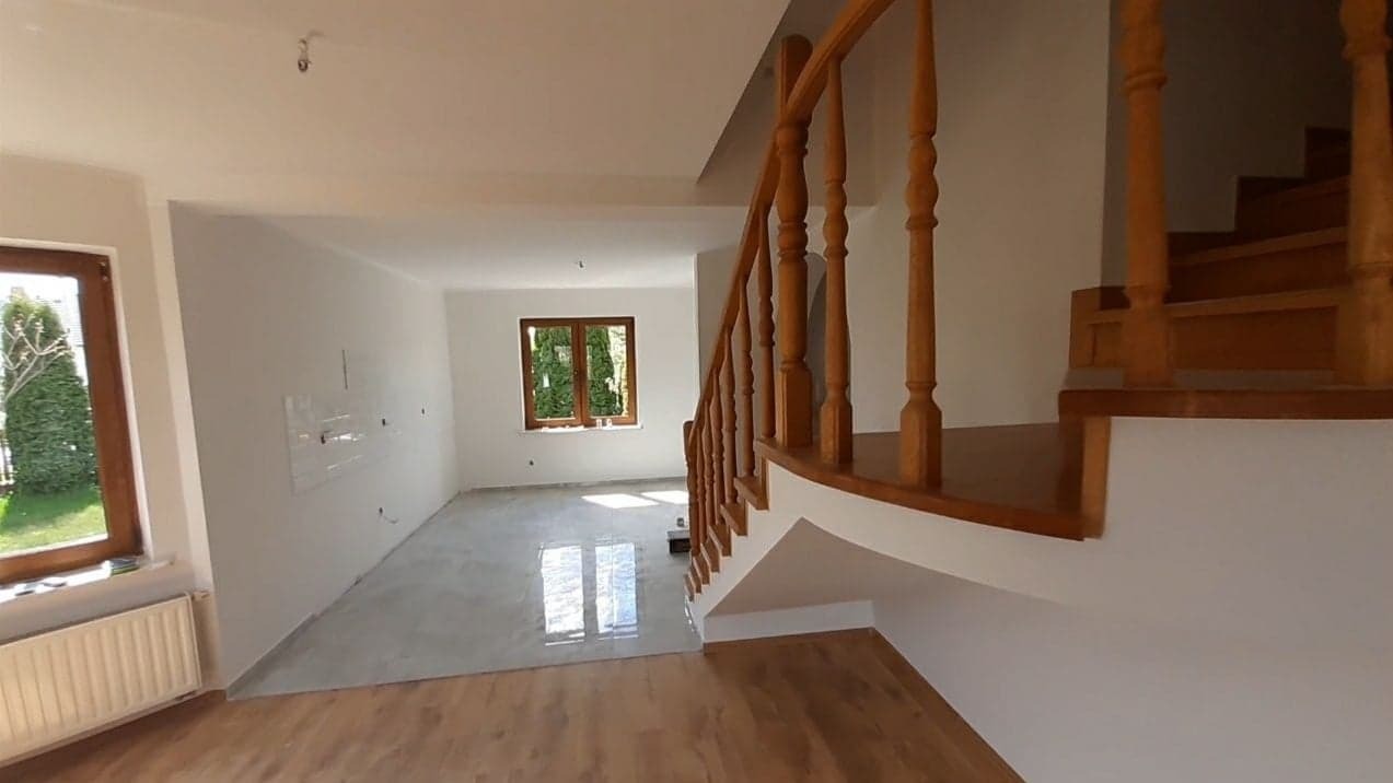 Wnętrze domu w trakcie prac wykończeniowych: świeżo pomalowane białe ściany, drewniane schody z balustradą i częściowo wylana posadzka samopoziomująca z widocznymi refleksami światła wpadającego...