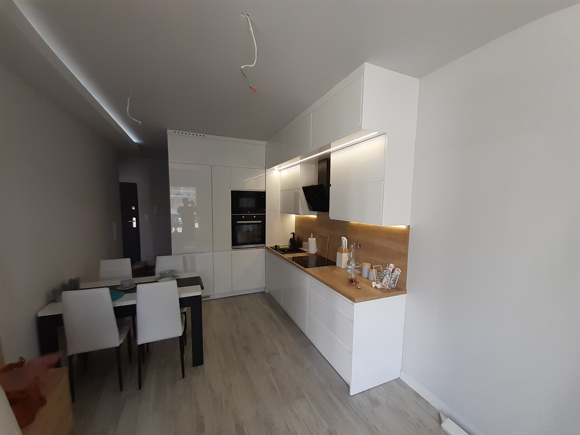 Nowoczesna kuchnia z białymi, lakierowanymi frontami, drewnianym blatem i oświetleniem LED pod szafkami, widoczny stół z krzesłami w tle, kabel zwisający z sufitu.