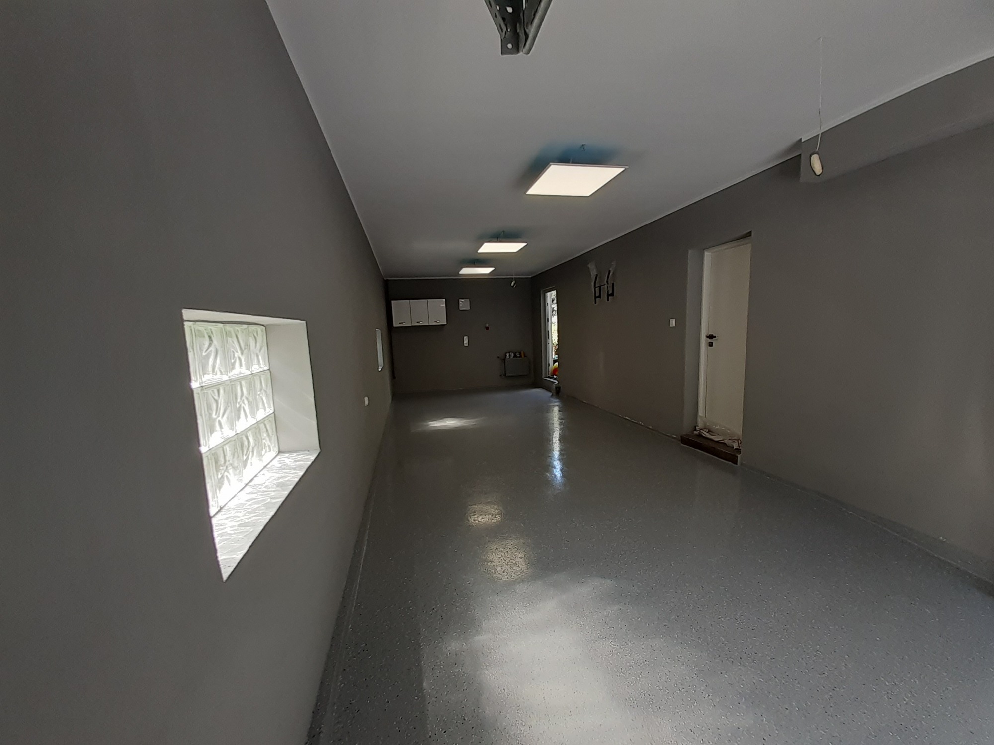 Wnętrze odnowionego korytarza z szarymi ścianami, błyszczącą podłogą z efektem lastryko, oświetleniem LED i oknem z luksferami.