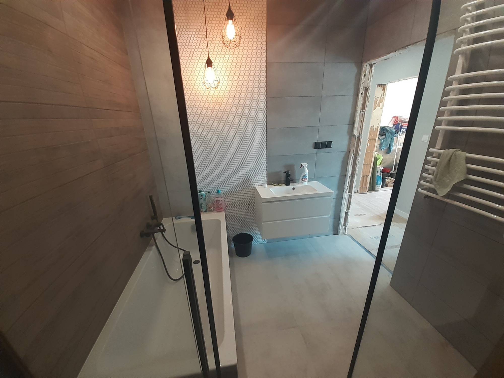 Widok na łazienkę w trakcie prac: drewnopodobne płytki na ścianie, biała mozaika, wisząca lampa z żarówką Edison, biała umywalka z szafką, kabina prysznicowa z czarnymi profilami, grzejnik...