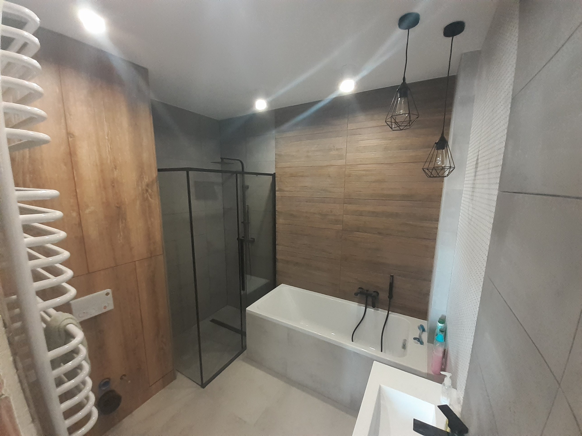 Nowoczesna łazienka z drewnopodobnymi płytkami na ścianie, kabiną prysznicową typu walk-in z czarnymi ramami, białą wanną z czarną armaturą, grzejnikiem drabinkowym i geometrycznymi lampami wiszącymi.