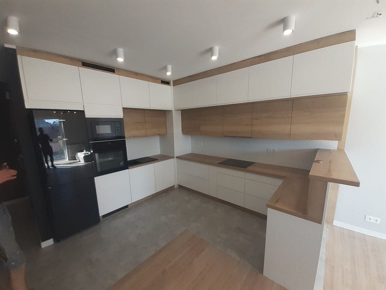 Nowoczesna kuchnia z białymi szafkami i drewnianymi akcentami, czarny sprzęt AGD, oświetlenie punktowe sufitowe, widok z poziomu podłogi.