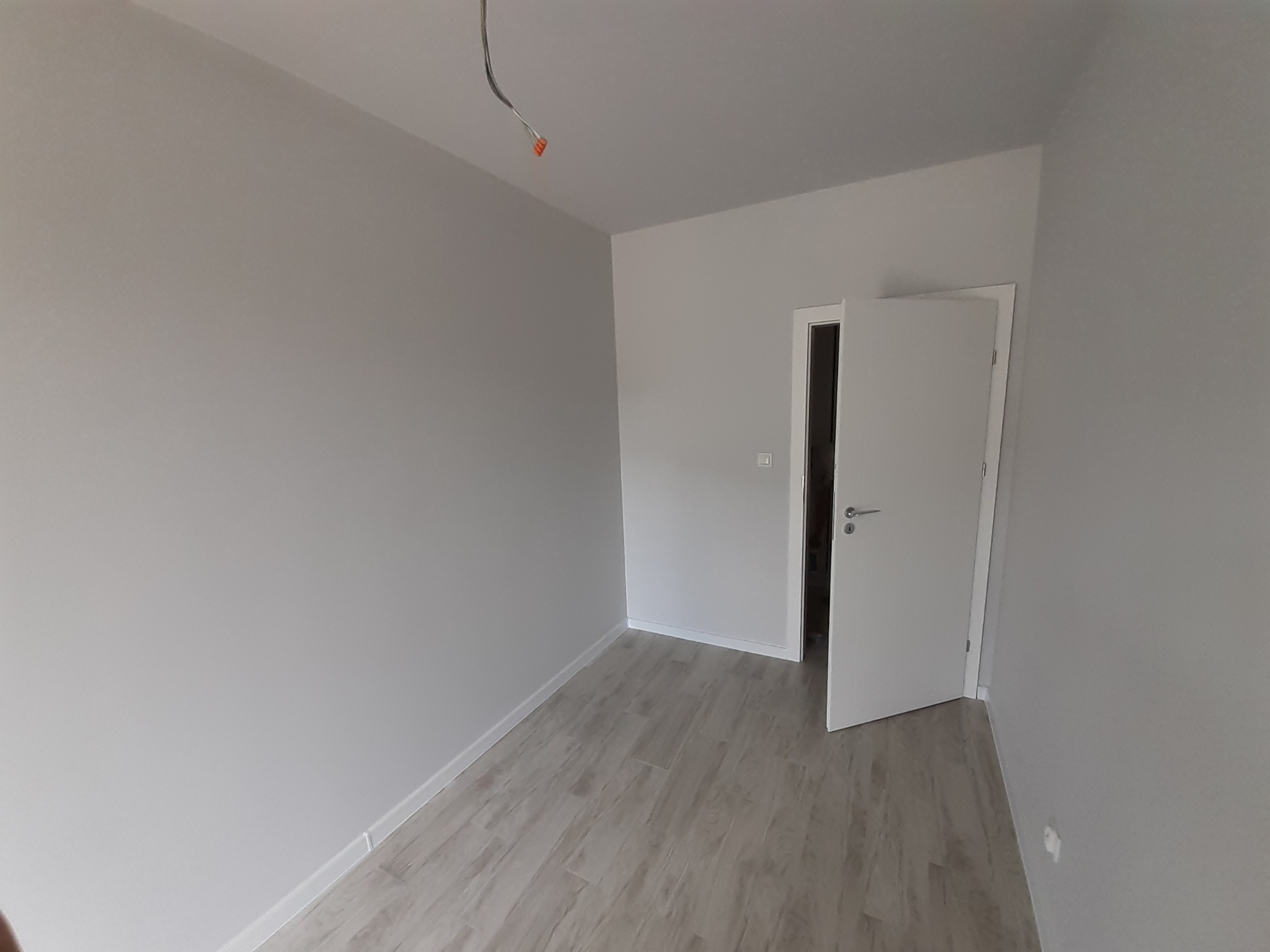 Pomalowane na szaro wnętrze z drewnopodobną podłogą, otwarte białe drzwi wewnętrzne, wystające kable ze ściany na suficie.