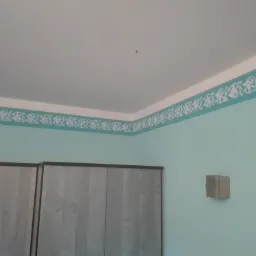 Turkusowa ściana z ornamentem malowanym wzdłuż sufitu, widoczny fragment szafy i wystający element na ścianie.