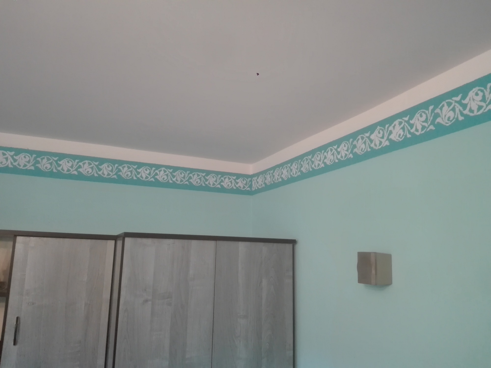 Turkusowa ściana z ornamentem malowanym wzdłuż sufitu, widoczny fragment szafy i wystający element na ścianie.