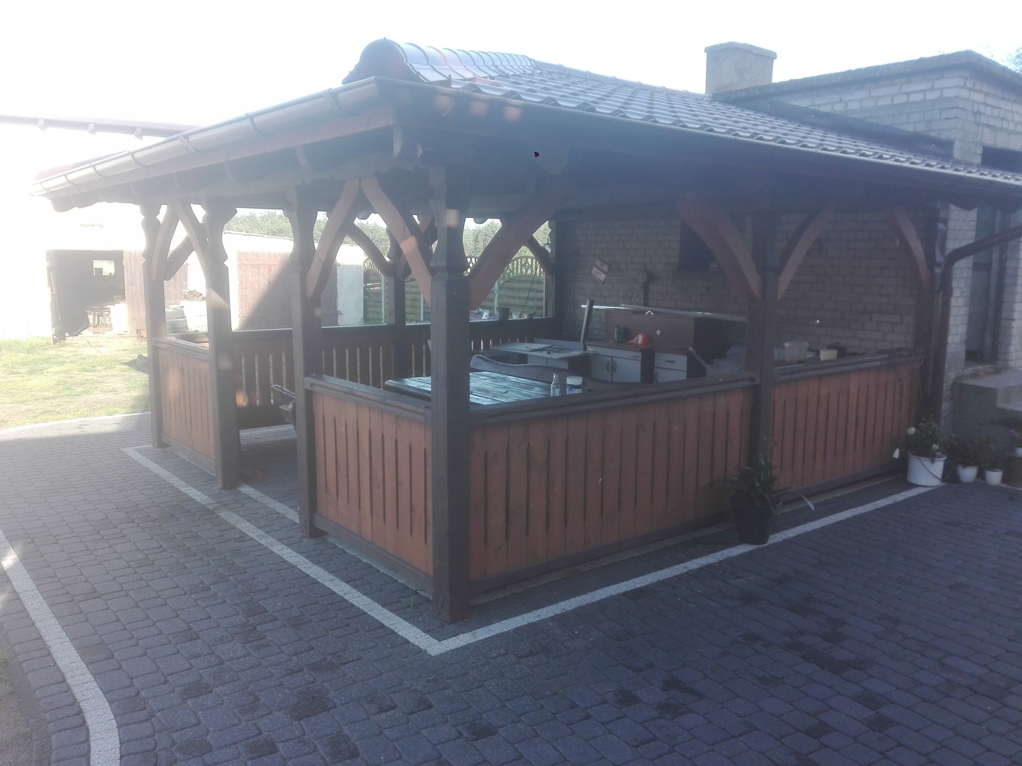 Zadaszona, drewniana altana z grillem murowanym w tle, otoczona brukowaną nawierzchnią.