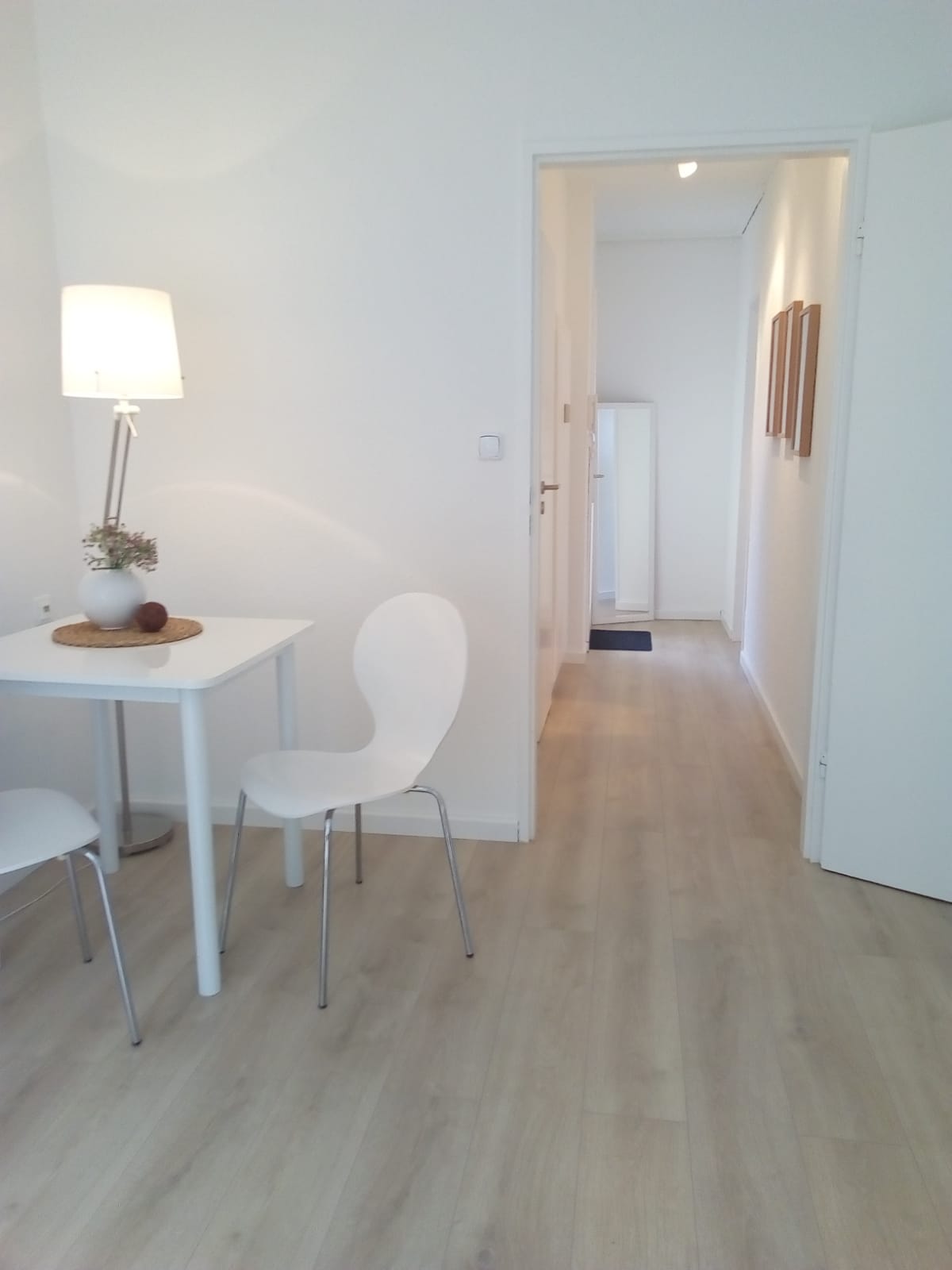 Jasne wnętrze mieszkania: parkiet w kolorze naturalnego drewna, białe ściany, minimalistyczny stół z lampą i dekoracją, białe krzesła oraz widok na korytarz z lustrem w tle.